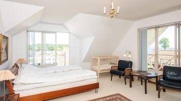 A bedroom with a large bed, a wooden headboard, a bedside table with a lamp, a small table with chairs, a crib, and a view through large windows.