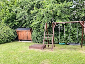 A wooden swing set with a ladder and a tire swing in a grassy yard with a shed and dense foliage in the background.