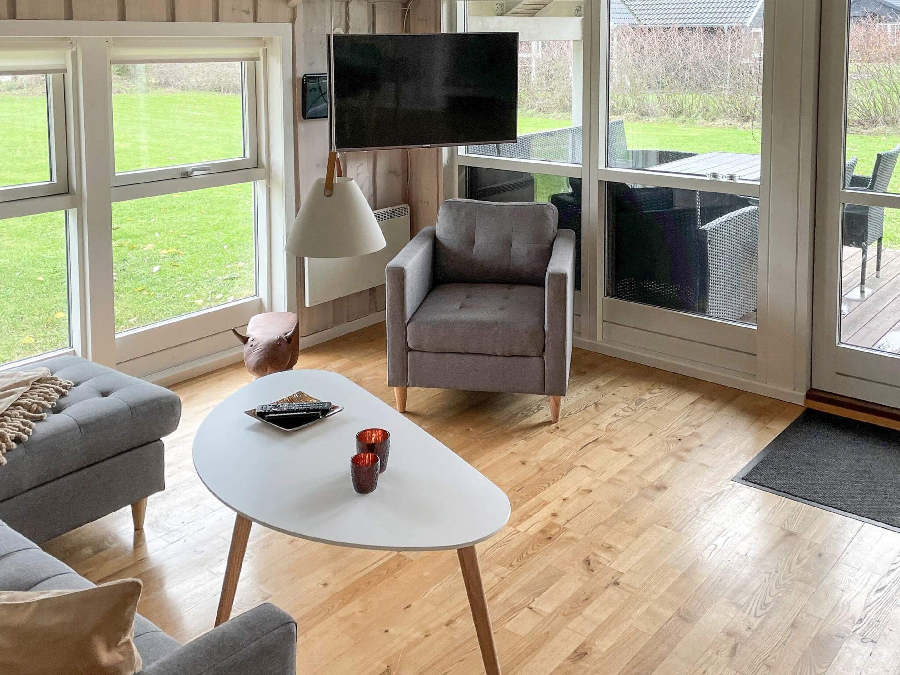 A modern living room with a grey sofa, a white coffee table, and a grey armchair. A flat-screen TV is mounted on the wall, and there is a large window providing a view of the outdoors.