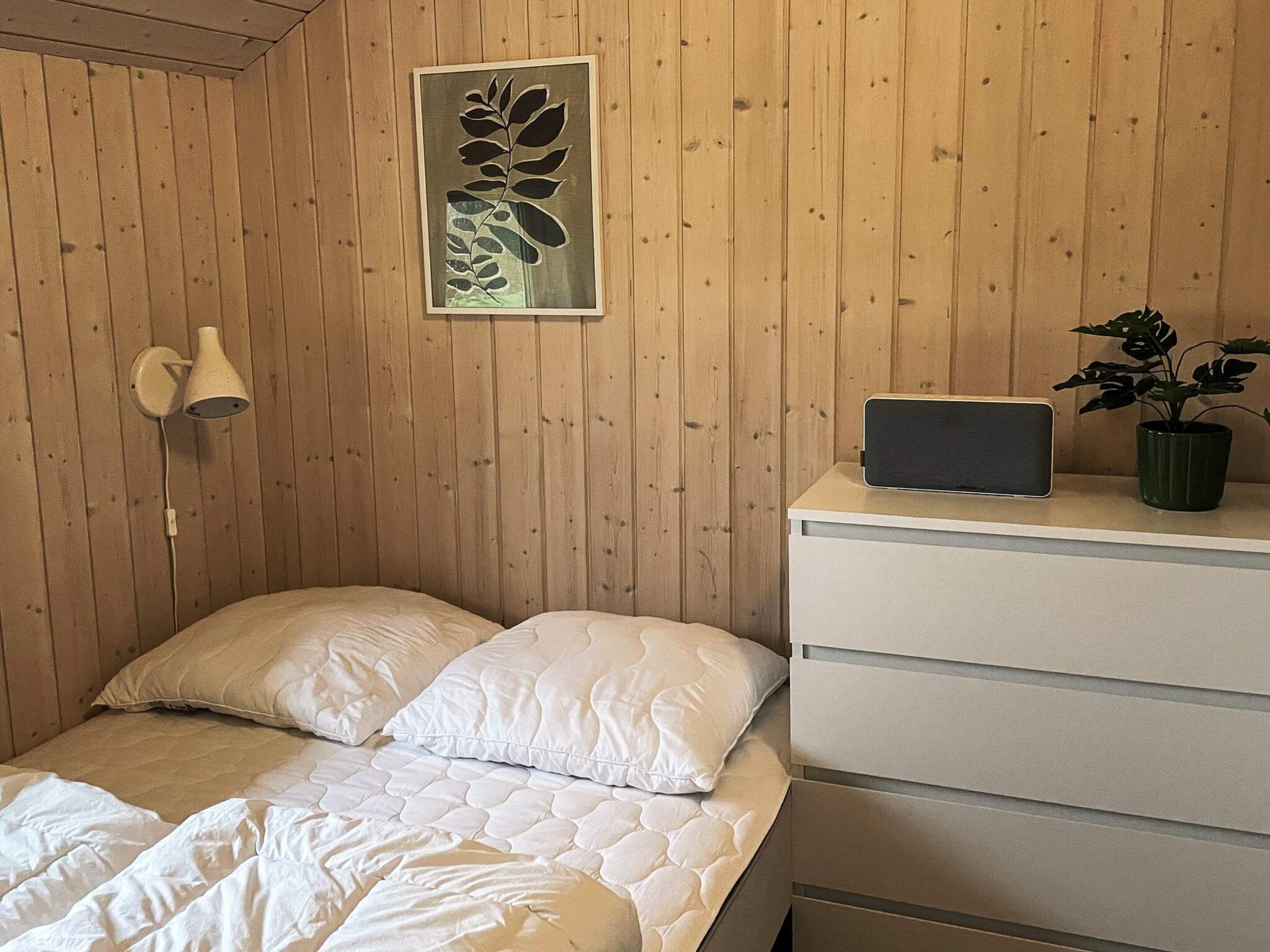 Et soveværelse med en seng, et natbord med en lampe, et billede i ramme og en potteplante.