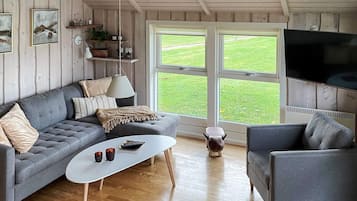 Un salón acogedor con un sofá gris, una mesa de centro blanca y un sillón gris. Hay un televisor montado en la pared y un ventanal con vistas a un césped verde.