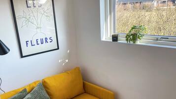 Ein Zimmer mit einem gelben Sofa, einem gerahmten Kunstwerk an der Wand und einem Fenster mit Blick auf ein Haus und Bäume.
