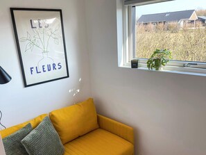 A room with a yellow sofa, a framed artwork on the wall, and a window with a view of a house and trees. - 6 Person Holiday Home in Sydals (Sydals)