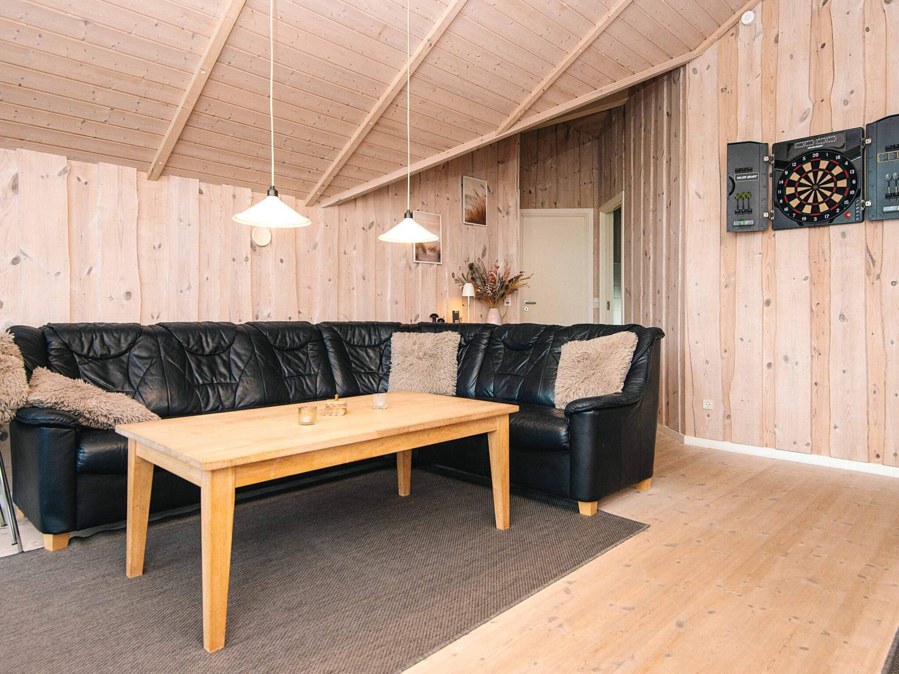 A room with a black leather sofa, a wooden table, hanging pendant lights, and a dartboard on the wall.