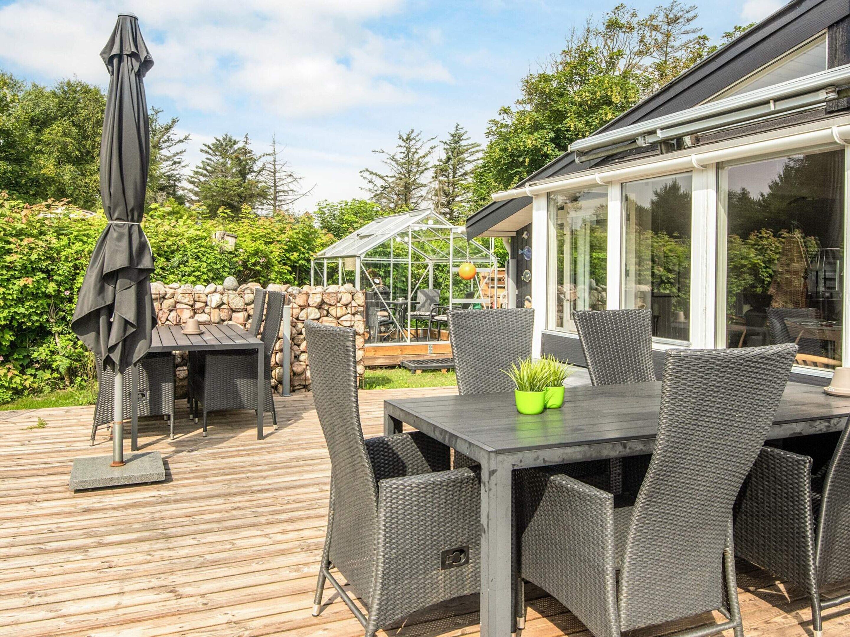 An outdoor patio with a table and chairs, a wooden deck, a stone wall, and a greenhouse.