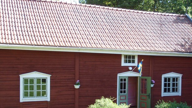 A red wooden building with a tiled roof, a green door, and a small flag. - 8 Person Holiday Home in Vimmerby (Vimmerby)