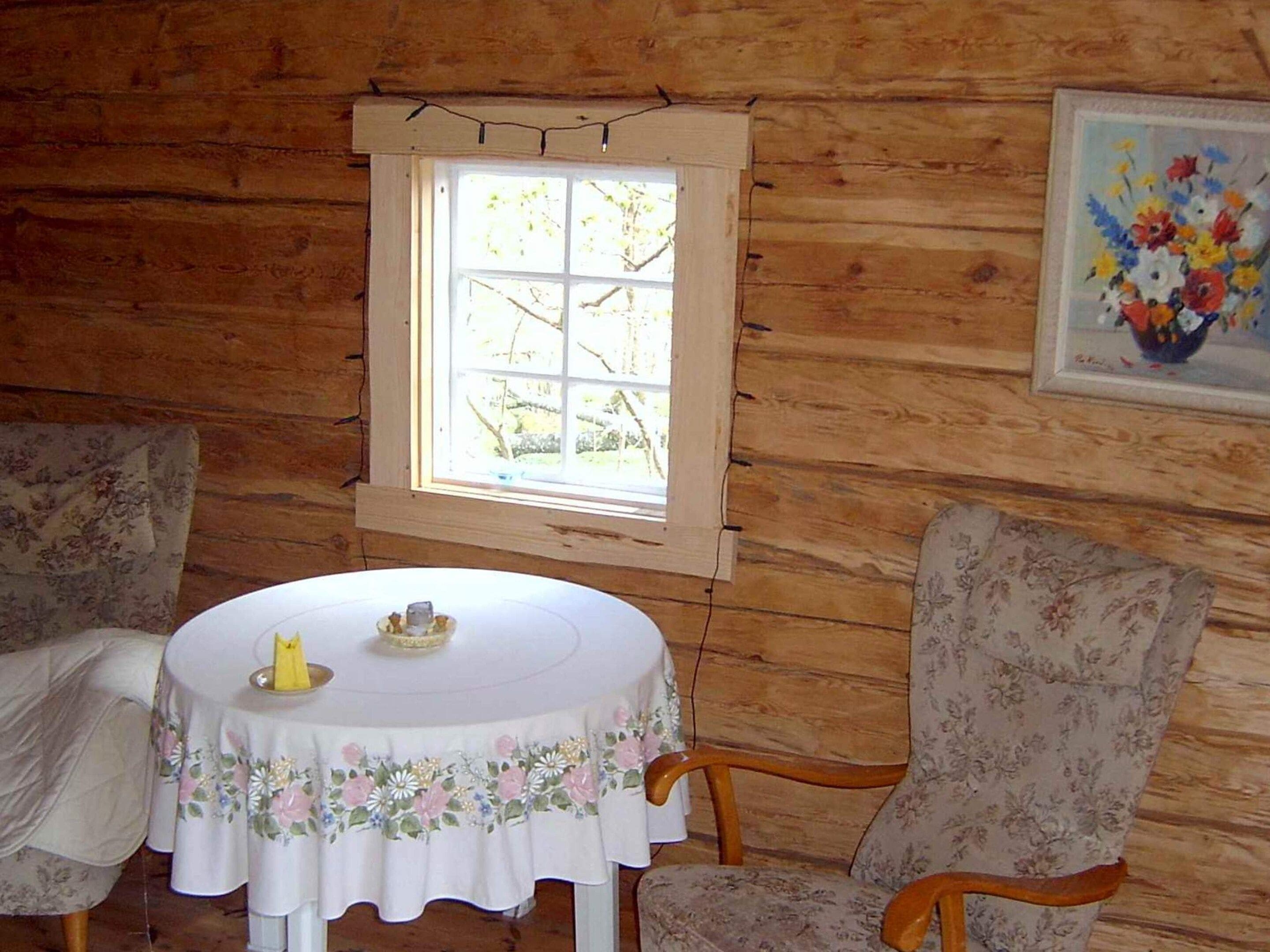 A wooden cabin interior with a round table, floral chairs, a window, and a framed floral painting.