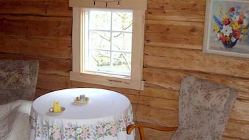 Interior de una cabaña de madera con una mesa redonda, sillas florales, una ventana y un cuadro con flores enmarcadas.