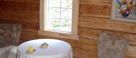 A wooden cabin interior with a round table, floral chairs, a window, and a framed floral painting.