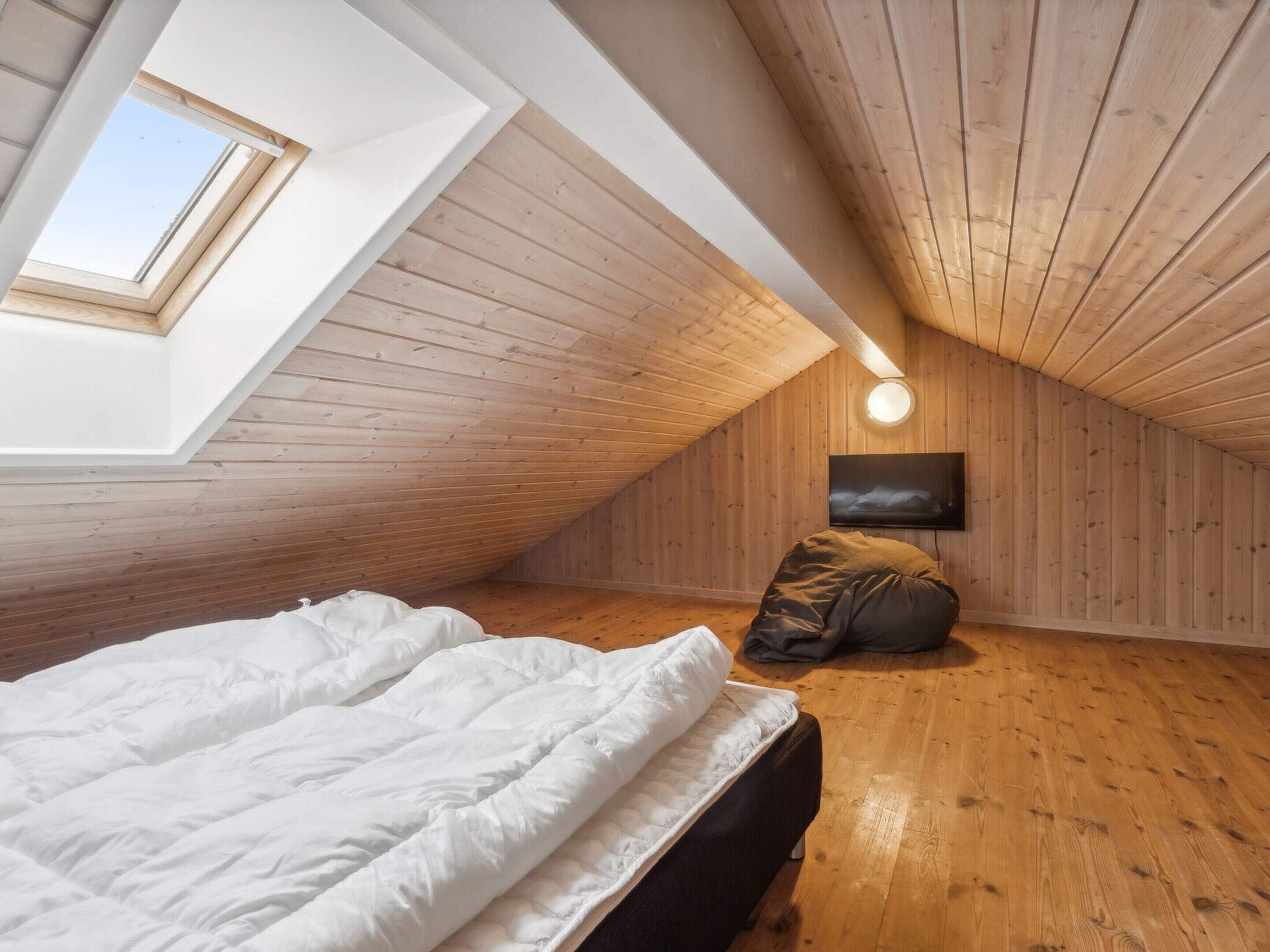 A bedroom with a sloped ceiling, wooden walls, a skylight, a bed, a pillow, and a wall-mounted light fixture.
