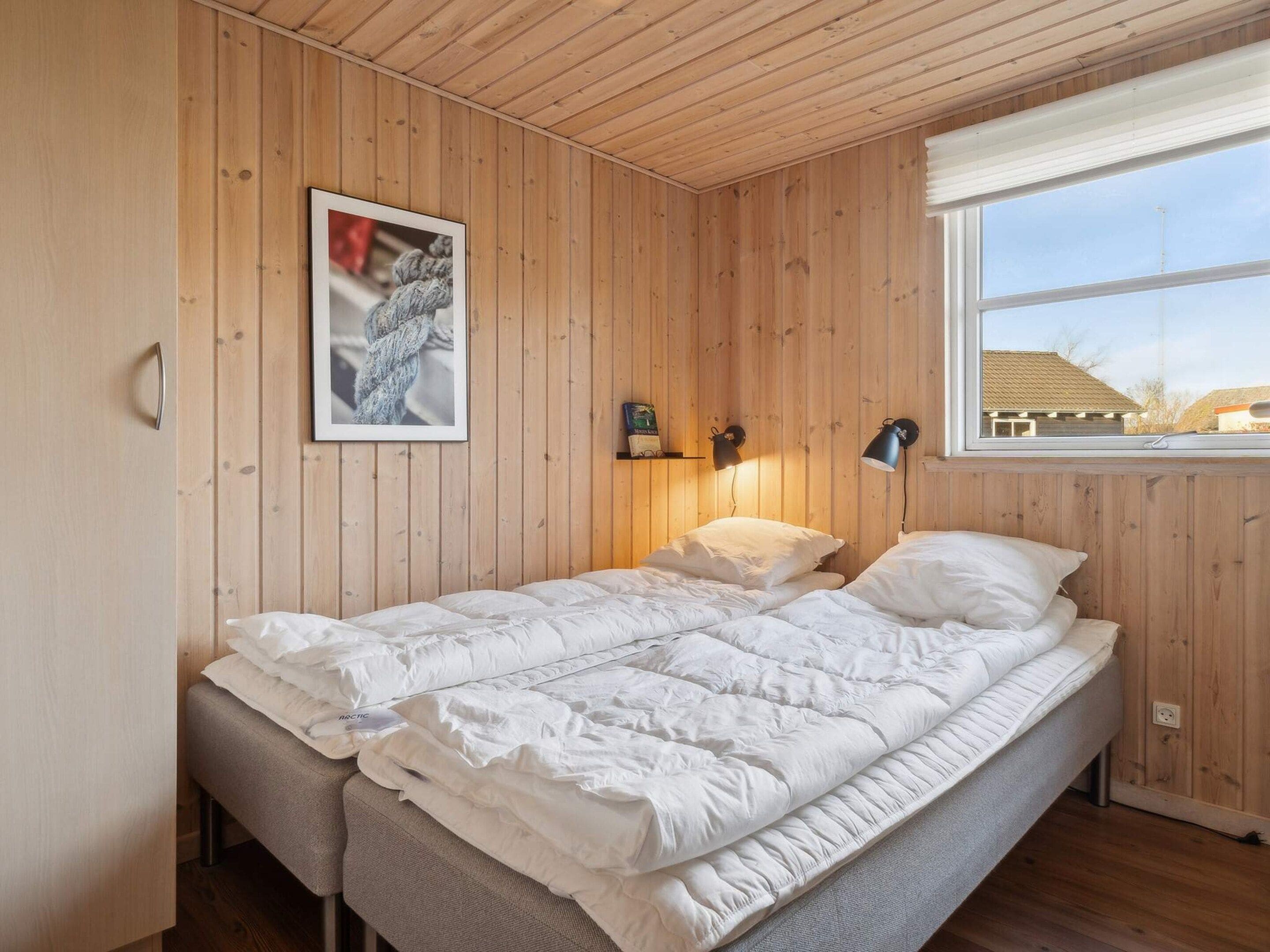 Un dormitorio con un revestimiento de madera, una cama con ropa de cama blanca, un cuadro enmarcado, dos lámparas fijadas a la pared y una ventana con vistas a edificios.