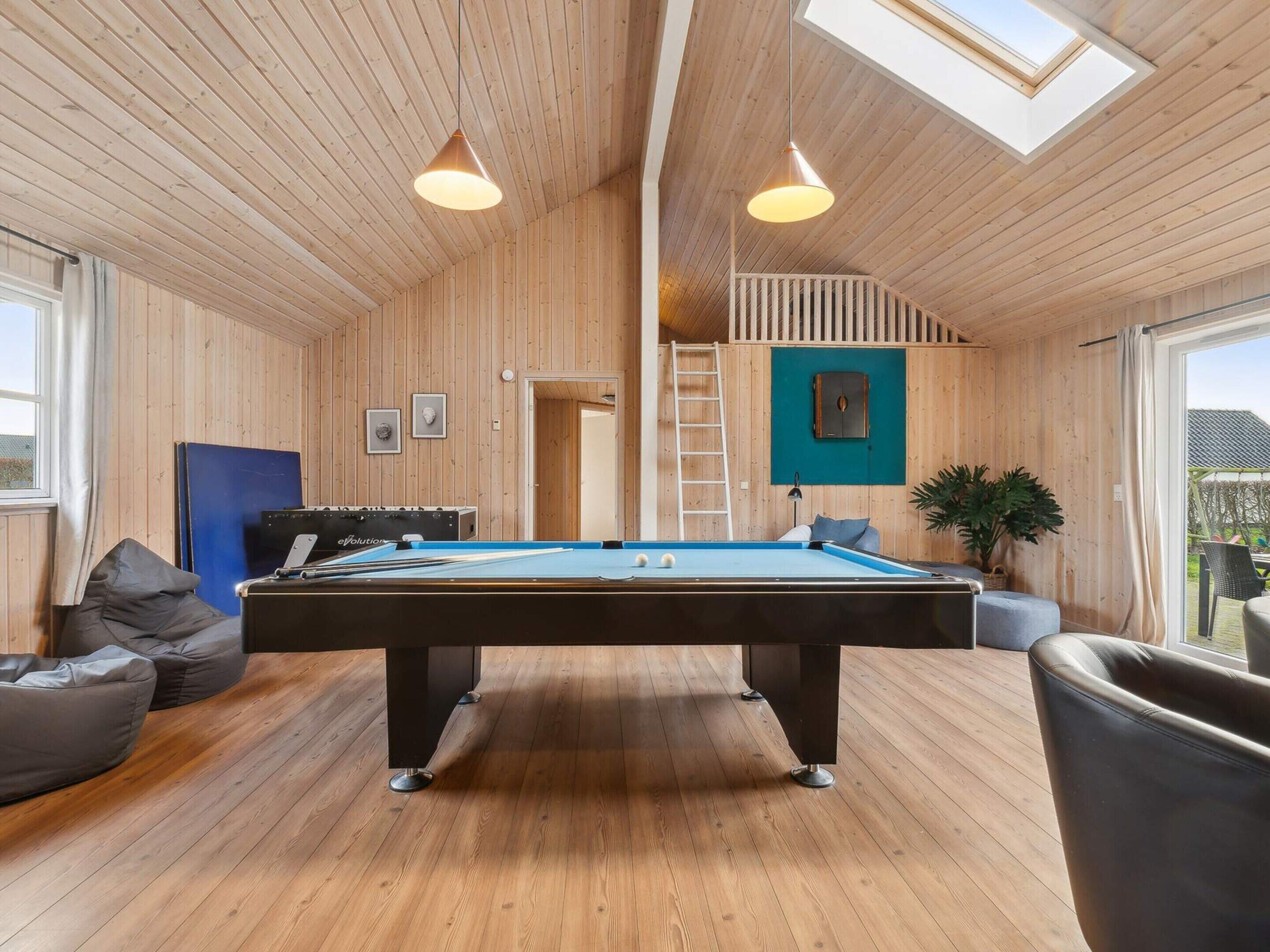 Une pièce en bois avec une table de billard, deux lampes suspendues, un canapé bleu et un escalier menant à une mezzanine.