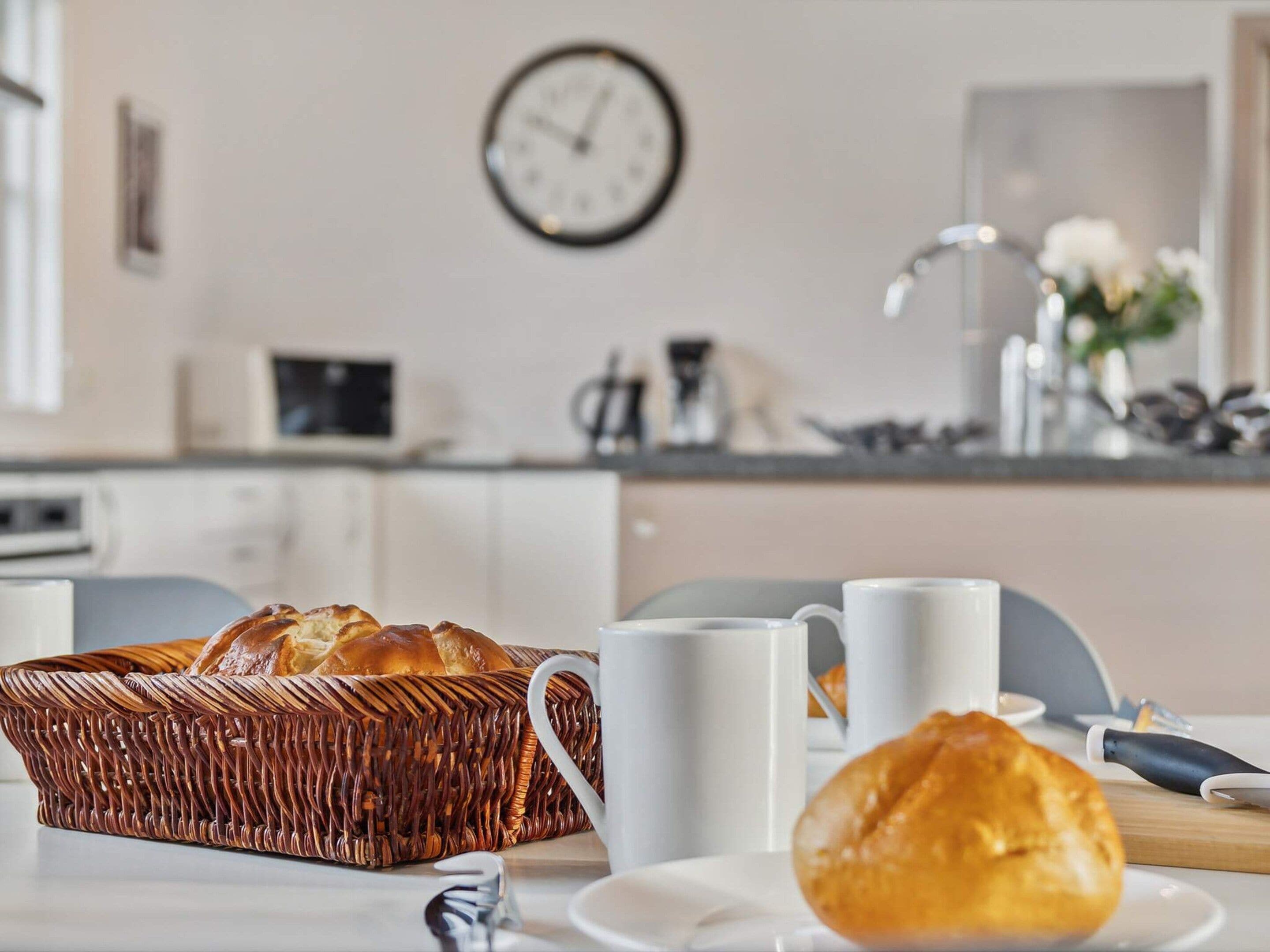 Une cuisine avec un panier à pain en osier, des tasses blanches et une horloge au mur.