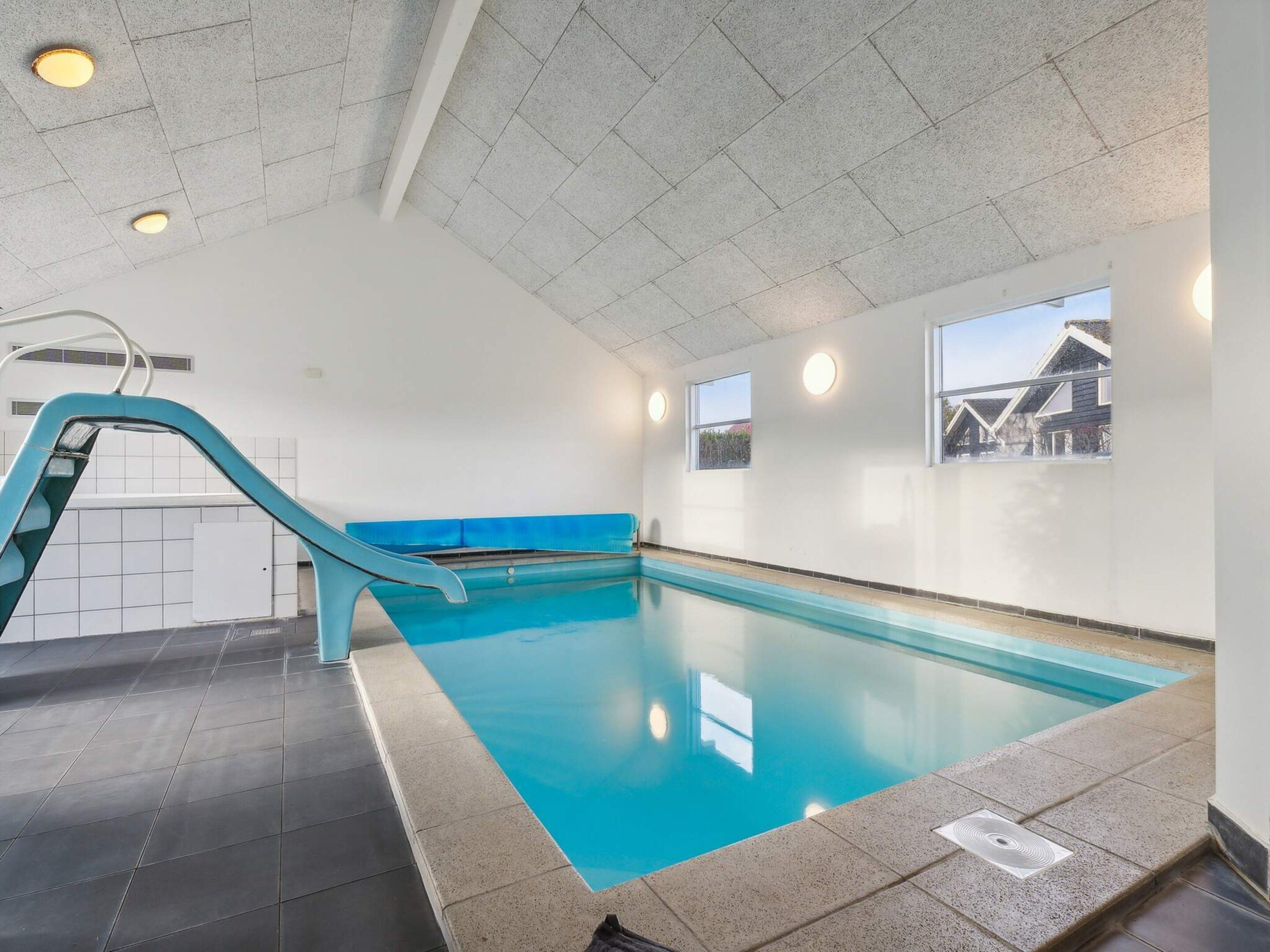 An indoor swimming pool with a blue slide, tiled walls, and windows with views of houses.