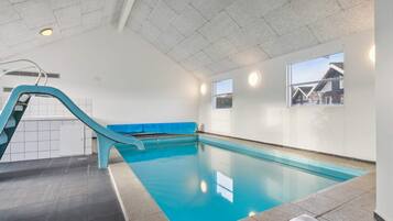 An indoor swimming pool with a blue slide, tiled walls, and windows with views of houses.
