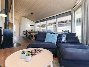 A modern living room with a dark blue sofa, a round coffee table with magazines and plants, and large windows with curtains.
