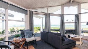 Ett modernt vardagsrum med ett stort fönster med utsikt över havet, en mörkgrå soffa, ett soffbord i trä och ett litet runt bord med en tidning på.