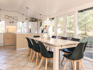 A modern kitchen with a central island, a dining area with a long table and chairs, and large windows offering a view of the outdoors. - 6 Person Holiday Home in Albaek (Aalbaek)