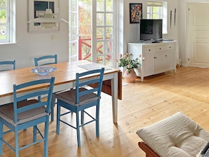 Un área de comedor con una mesa de madera y sillas azules, un armario blanco con un televisor, y una ventana con vista a follaje.