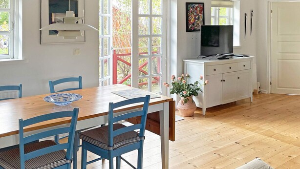 A dining area with a wooden table and blue chairs, a white cabinet with a television, and a window with a view of greenery. - Holiday Home in SÃ¶lvesborg (Sölvesborg)