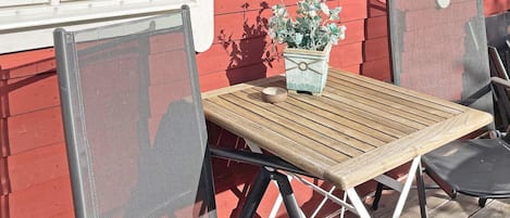 An outdoor seating area with a table and chairs, a potted plant, and a window with green shutters.