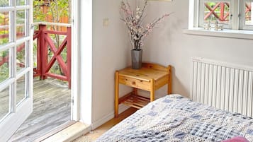 Un dormitorio con cama, mesita de noche de madera, un florero con ramas, ventana con vista a una estructura roja y un radiador.