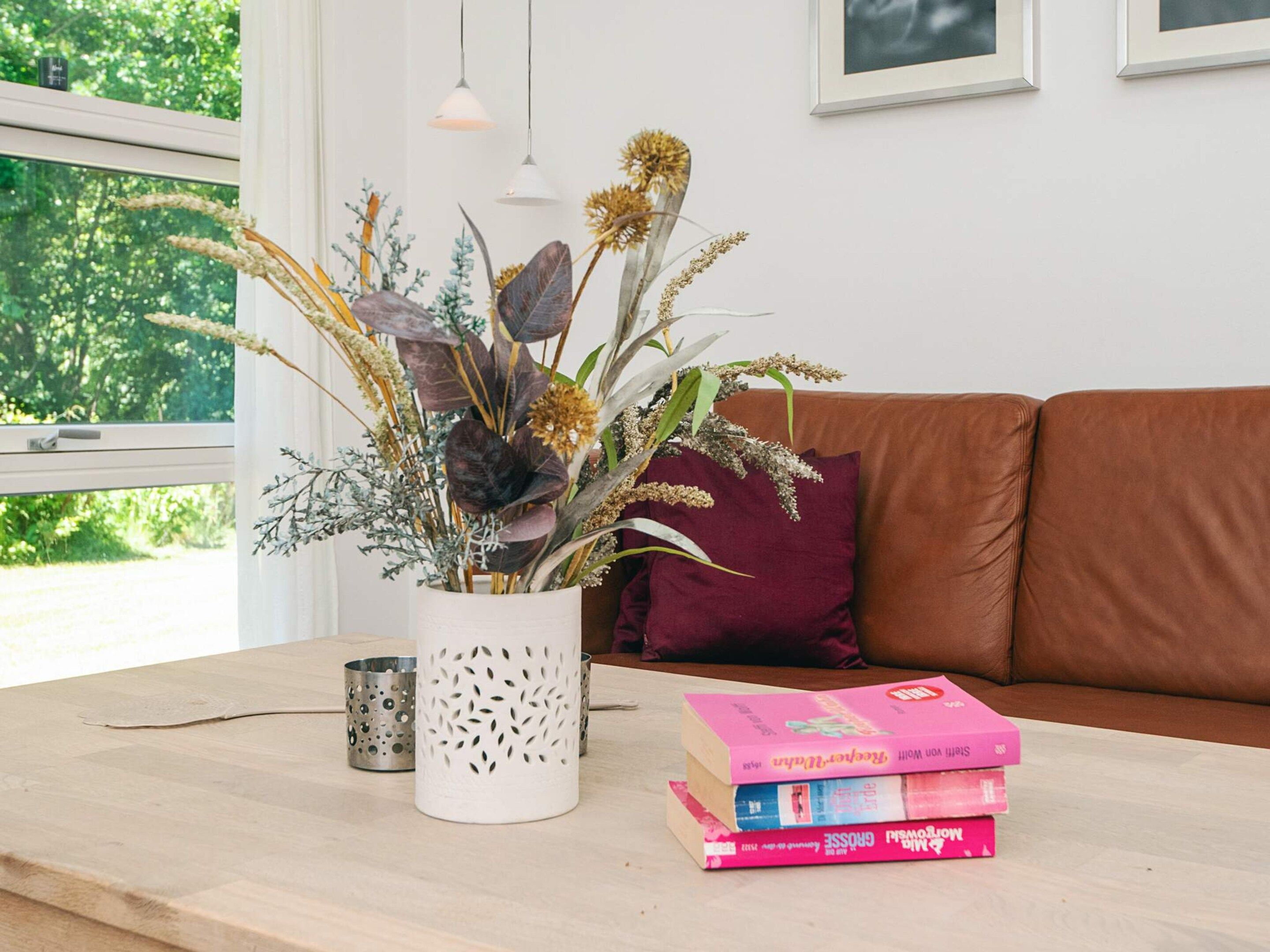 A room with a wooden table, a white vase with dried plants, a brown leather couch, and a stack of books.