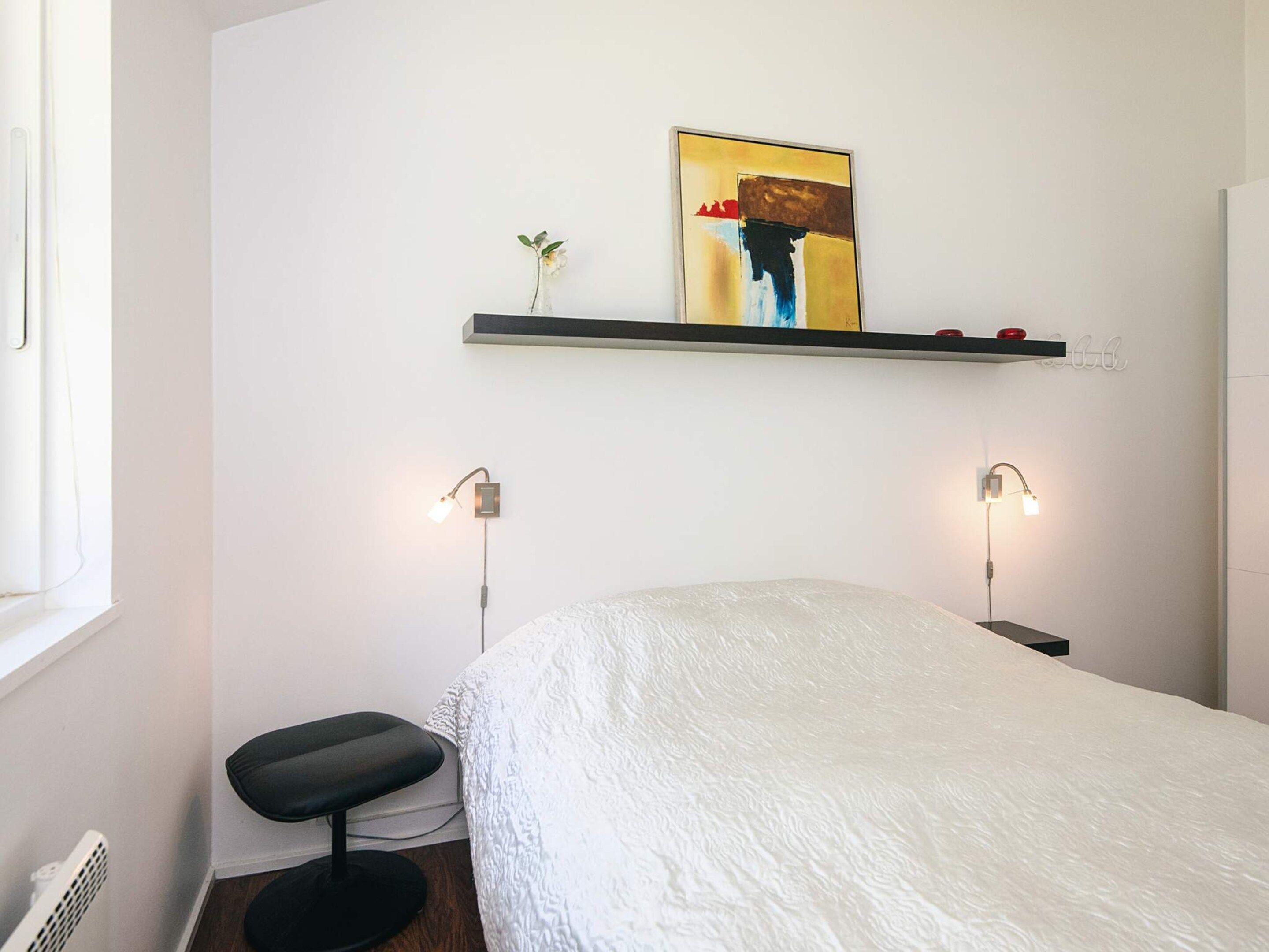 Une chambre à coucher avec un lit, un petit tabouret, une étagère fixée au mur sur laquelle est posée une peinture, et deux lampes de chevet.