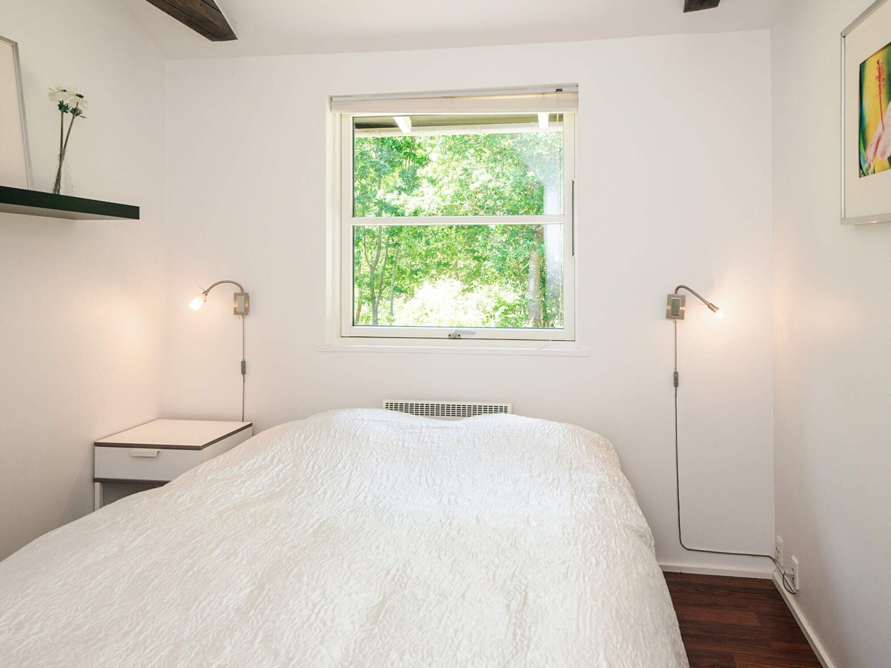 A bedroom with a large bed, a window with a view of trees, a bedside table, and a wall-mounted lamp.