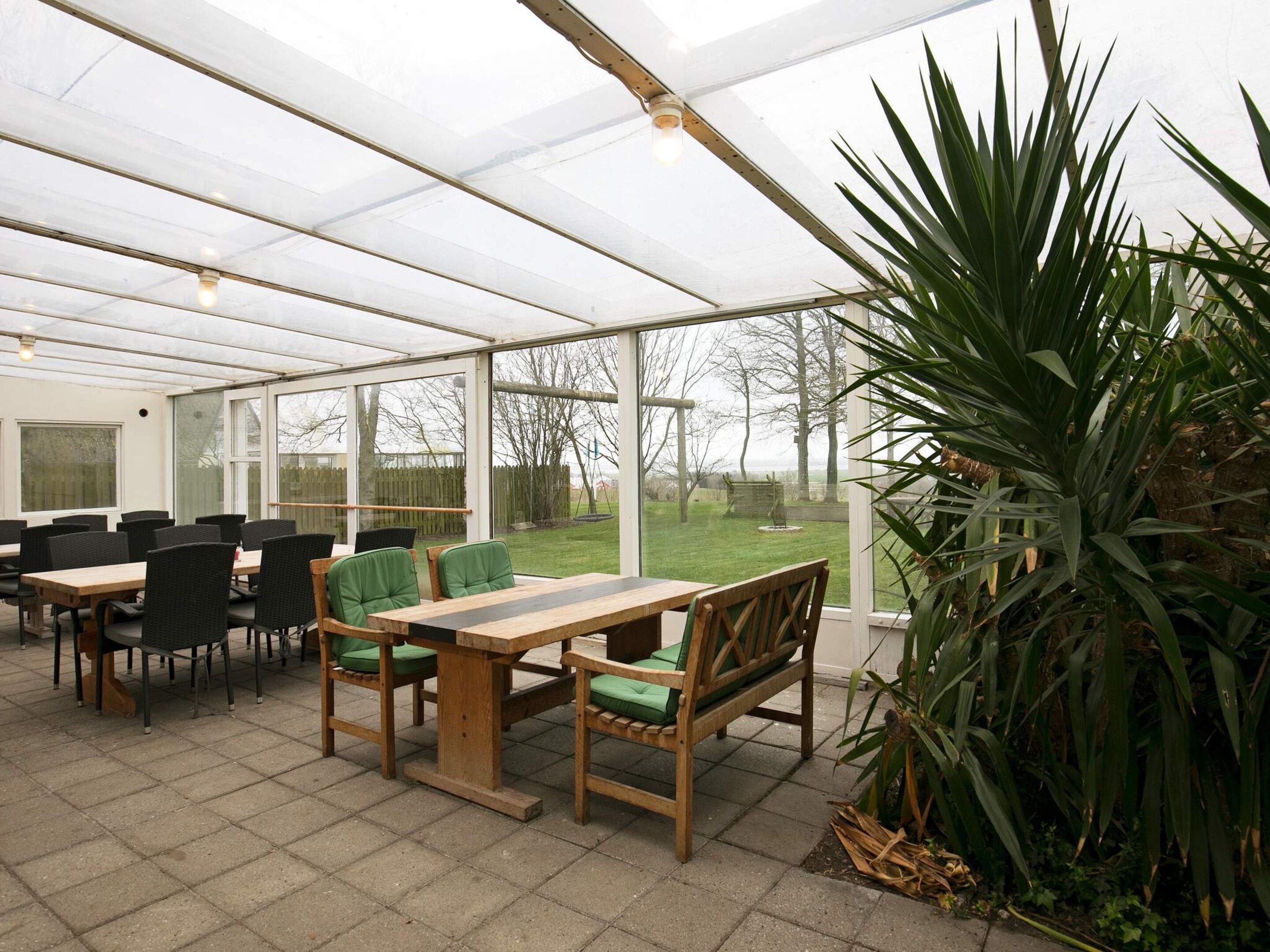 Un invernadero con una mesa y sillas de madera, ventanas amplias y vistas al exterior.
