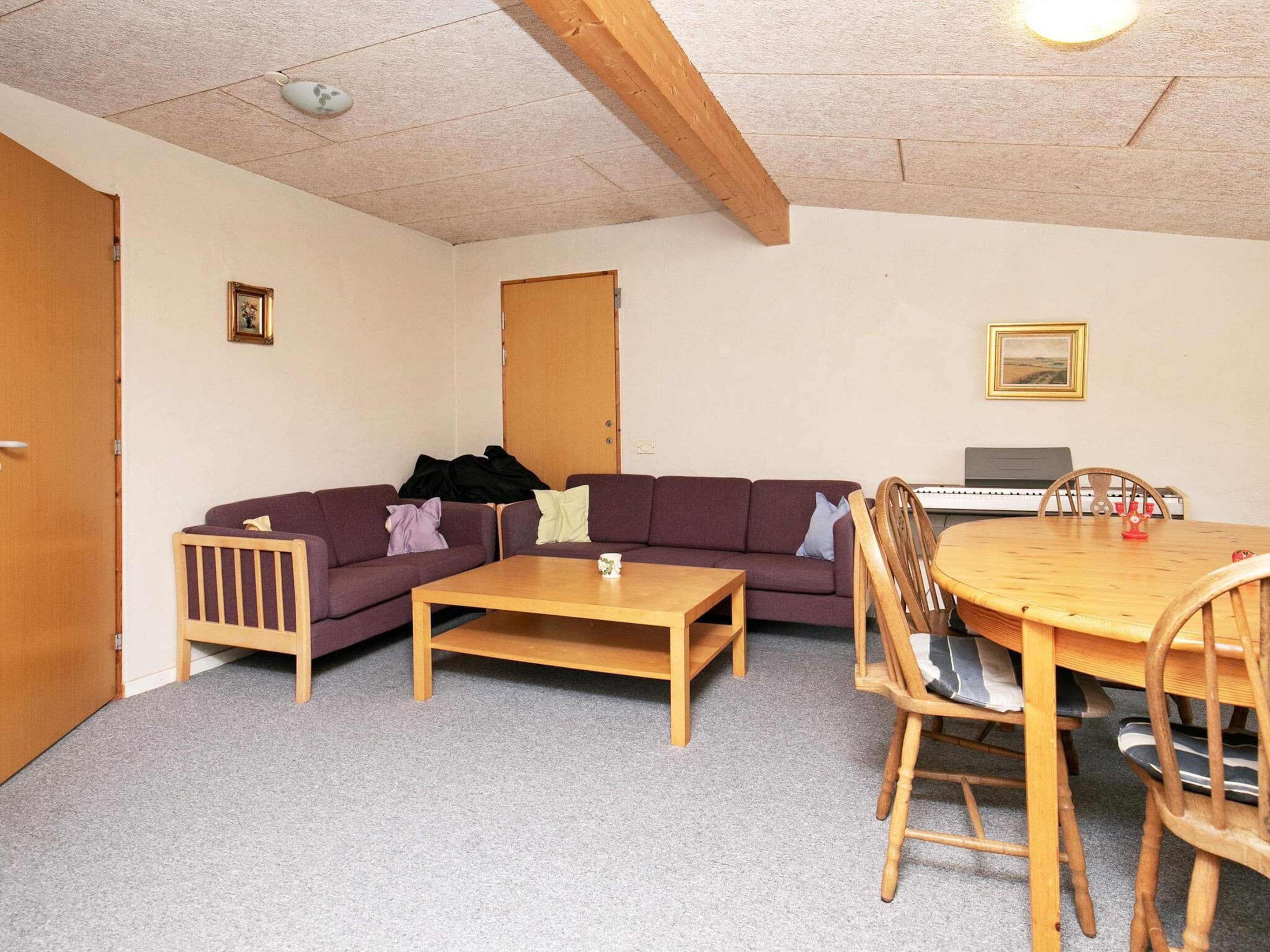 Een kamer met een houten plafond, een bank, een salontafel, een eettafel met stoelen, een toetsenbord en een lijst met een afbeelding aan de muur.