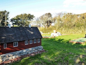 Een klassiek rood huis met een stenen fundering en rieten dak, omgeven door een verzorgd gazon en bomen.