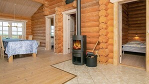 A log cabin interior with a central wood-burning stove, a dining area with a table and chairs, and a bedroom with a bed. - 5 Star Holiday Home in Frostrup (Frøstrup)