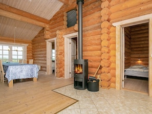 A log cabin interior with a central wood-burning stove, a dining area with a table and chairs, and a bedroom with a bed. - 5 Star Holiday Home in Frostrup (Frøstrup)