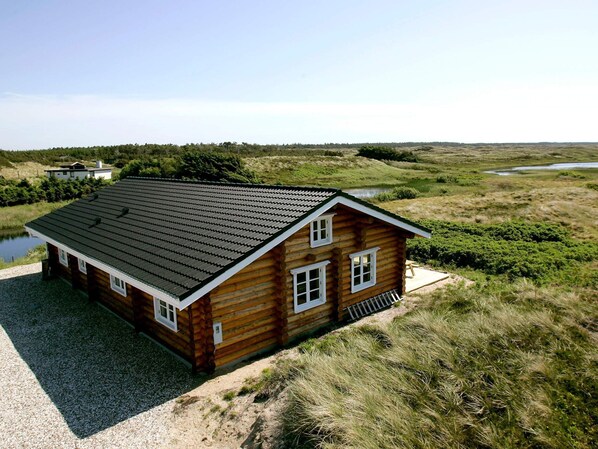 A log cabin with a sloped roof, a gravel driveway, and a pond in the background.
