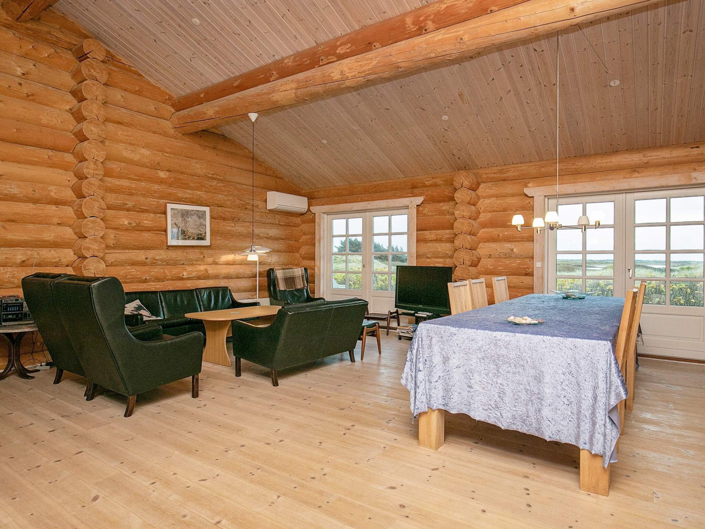 A log cabin interior with a dining table, two green leather sofas, a television, and a large window offering a view of the outdoors.