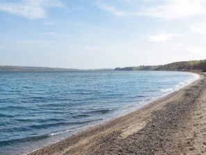 Ein Kiesstrand mit ruhiger See, blauem Himmel und fernen Hügeln.