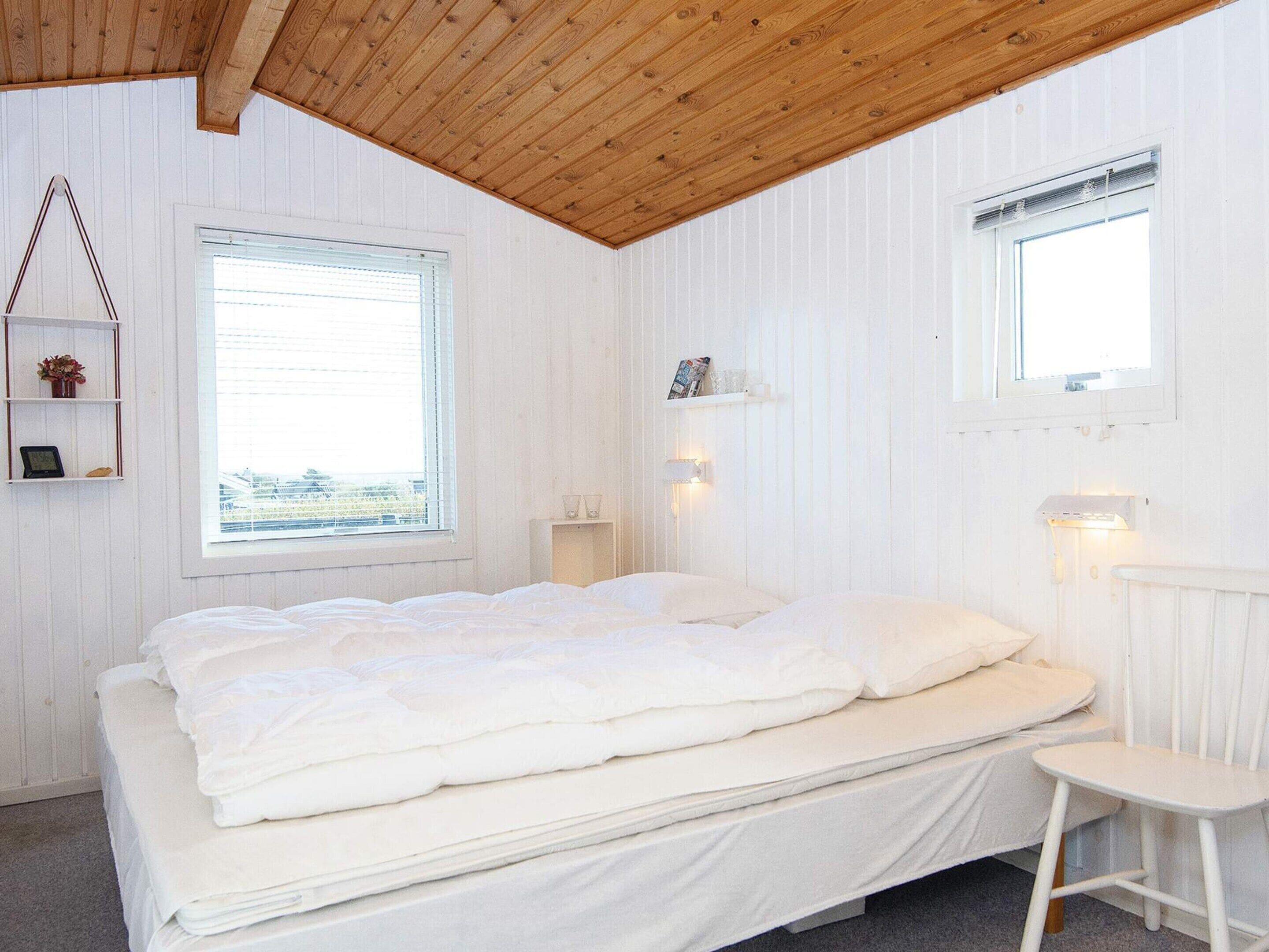 A bedroom with a wooden ceiling, a bed with white bedding, a window, and a small bedside table.