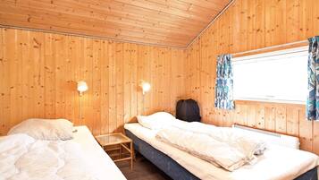 A room with two beds, wooden walls, a window with curtains, and two wall-mounted lamps.