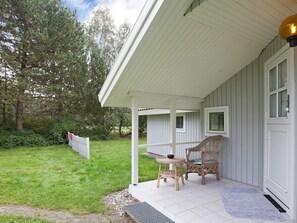 A covered porch with two wicker chairs and a small table, a white door, and a grassy yard with a fence.