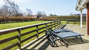 Eine Holzterrasse mit einer Liege aus Metall und einem kleinen Tisch, mit Blick auf eine Rasenfläche mit Bäumen und einem Gebäude im Hintergrund.