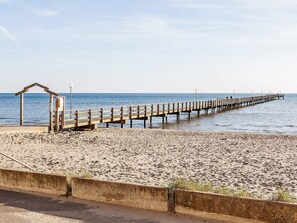 En strand med en träbrygga som sträcker sig ut i havet, ett litet skydd och en betongbarriär.
