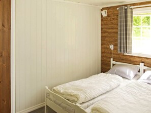 Un lit simple avec une literie blanche, une tête de lit en bois, une fenêtre avec des rideaux et un mur avec un lambris vertical.