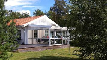 En vit byggnad med ett rött tegeltak, en inglasad veranda med stolar och en gräsklädd trädgård.