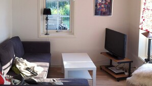 A living room with a sofa, a television on a stand, a small table, and a window with a view of greenery. - Holiday Home in Ronneby (Ronneby)