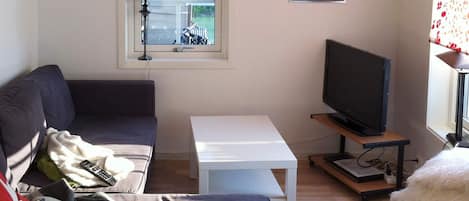 A living room with a sofa, a television on a stand, a small table, and a window with a view of greenery.