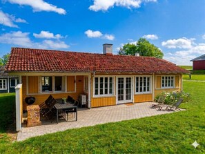 Una casa de madera amarilla con techo de tejas, un porche cubierto con mobiliario exterior y un patio con césped.