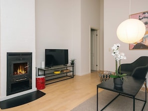 Uma sala de estar com lareira, televisão sobre um suporte preto, uma mesa com planta em vaso e uma porta que dá para outro cômodo.