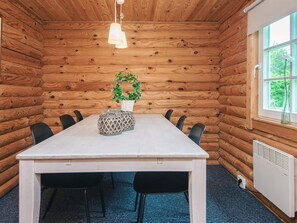 A wooden cabin interior with a white table, black chairs, a potted plant, and a window with a view of greenery.