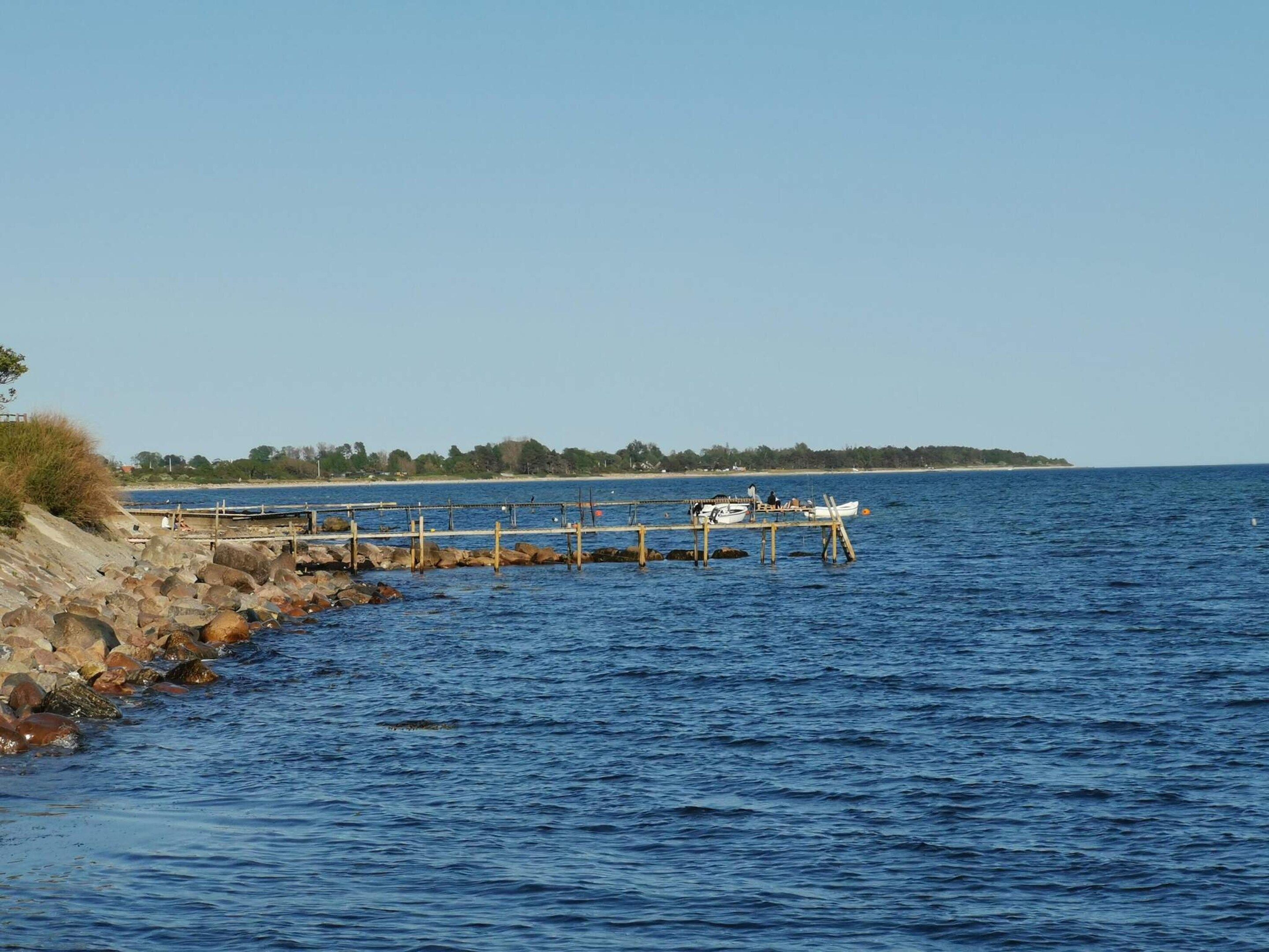 A rocky shoreline with a pier extending into calm waters, boats docked, and a distant tree-lined landmass.